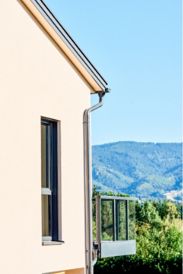 Außenansicht einer modernen Hausfassade mit Regenrinne, Fenster und Balkongeländer vor Bergkulisse.
