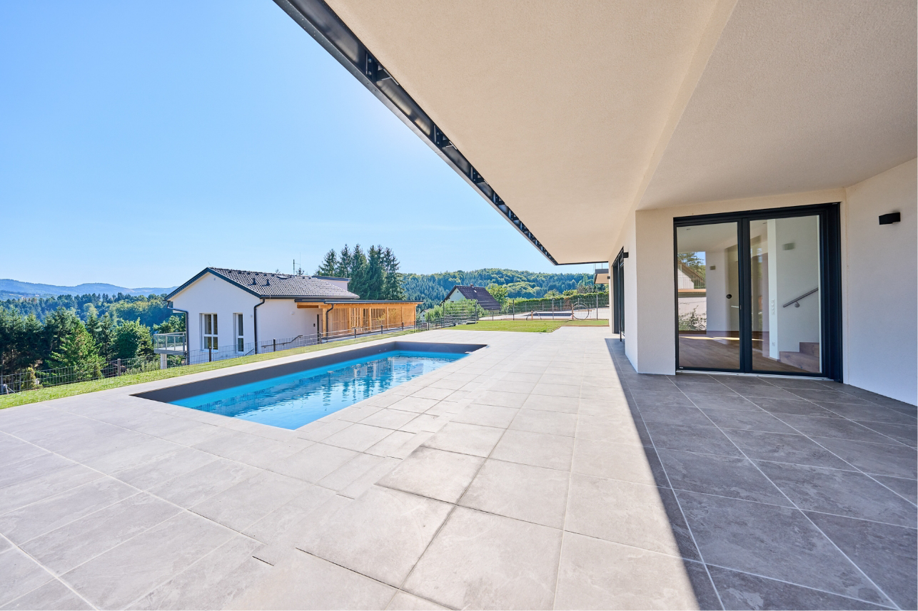 Große überdachte Terrasse mit Pool, hellem Fliesenboden und Panoramablick ins Grüne.